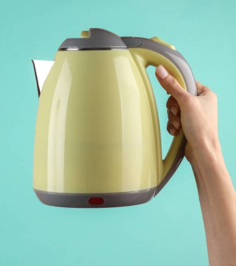 Kadının eli mavi arka planda elektrikli su ısıtıcısını tutuyor.