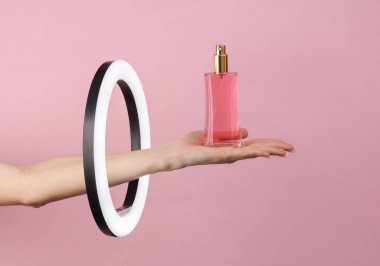 Kadının eli, pembe arka plandaki lambanın içinden parfüm şişesini tutuyor. Yaratıcı bir fikir. Güzellik ve moda konsepti