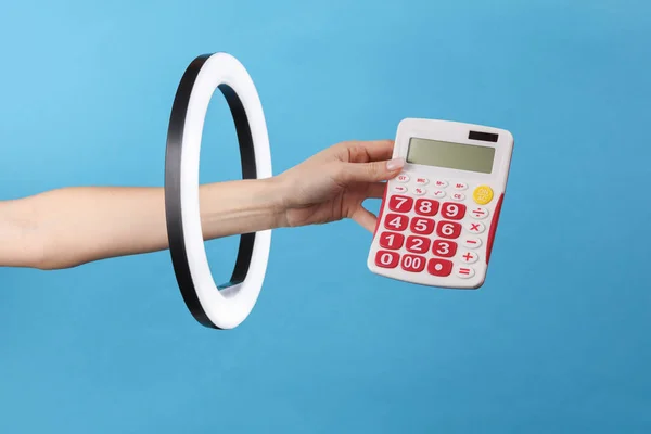 A woman\'s hand holds a calculator through a led ring lamp on a blue background. Creative idea. Business, economy concept