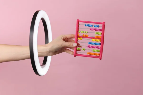 Woman\'s hand holds abacus through led ring lamp on pink background. Creative idea