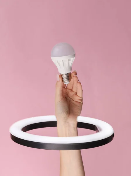 Woman\'s hand holds light bulb through led ring lamp on pink background. Creative idea.