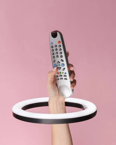 Woman\'s hand holds tv remote through led ring lamp on pink background. Creative idea.