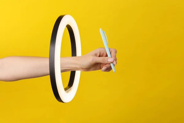 Woman's hand holds pen through led ring lamp on yellow background. Creative idea. Fashion concept