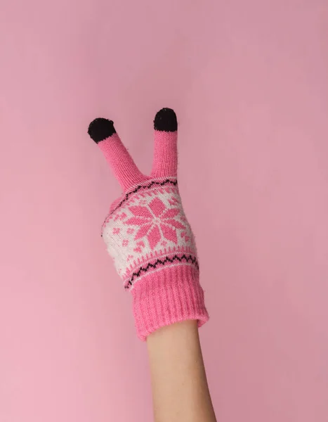 Women\'s hand in warm knitted glove with pattern showing v symbol of peace on a pink background