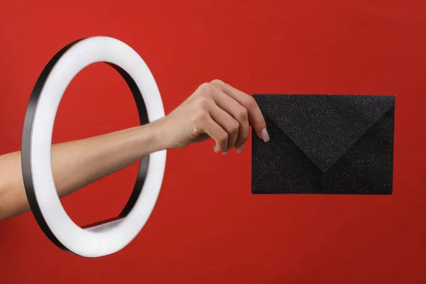 Woman\'s hand holds envelope through led ring lamp on red background. Creative idea.