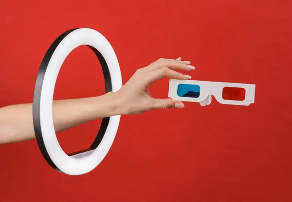 Woman\'s hand holds 3d glasses through led ring lamp on red background. Creative idea.