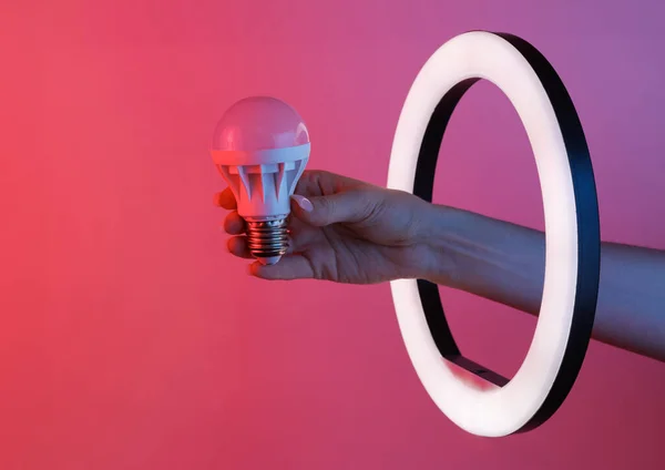 Woman\'s hand holds led light bulb through led ring lamp in red blue neon gradient light. Creative idea.
