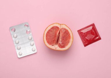 Contraception, protected sex concept. Half of a grapefruit symbolizing the female vagina, birth control pills with a condom package on a pink background
