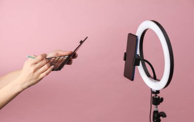 Güzellik blogu. Makyaj fırçası ve göz farı tutan kadın, pembe arka planda lamba ile akıllı telefon kamerasına yayın yapıyor.