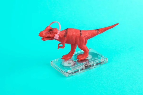 Toy red dinosaur tyrannosaurus rex with headphones and audio cassette on a turquoise background. Minimalism creative layout. Music lover