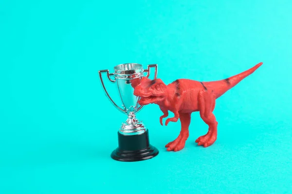 Toy red dinosaur tyrannosaurus rex with winner cup on turquoise background. Minimalism creative layout