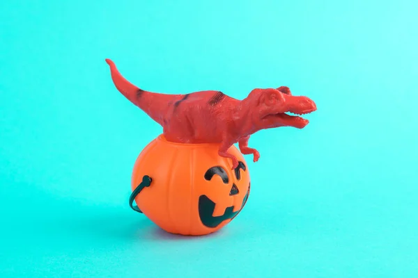 Toy red dinosaur tyrannosaurus rex with halloween bucket on a turquoise background. Minimalism creative layout. Trick or treat