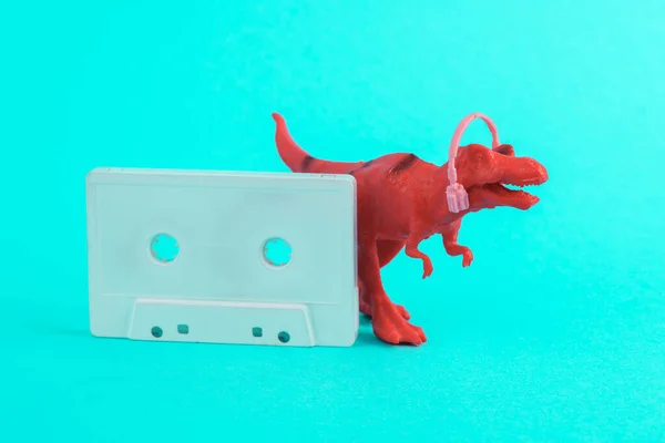 Toy red dinosaur tyrannosaurus rex with headphones and audio cassette on a turquoise background. Minimalism creative layout. Music lover. Retro 80s