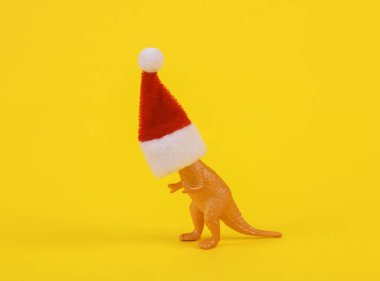 Sarı arka planda Noel Baba şapkası olan oyuncak dinozor. Yaratıcı Noel düzeni