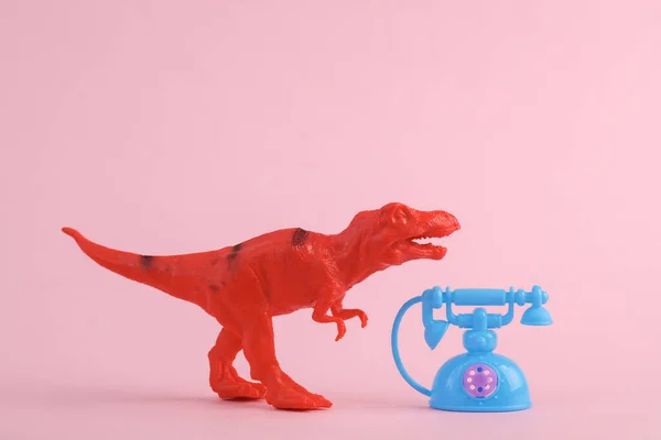 Toy red dinosaur tyrannosaurus rex with retro rotary phone on pink background. Minimalism creative layout