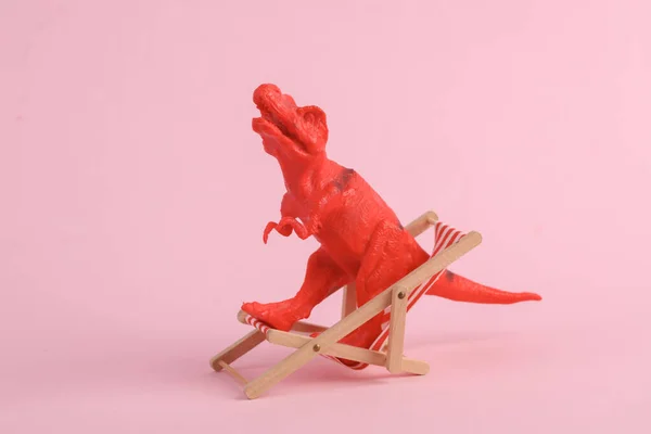 Toy red dinosaur tyrannosaurus rex with deck chair on pink background. Minimalism creative layout. Summer rest