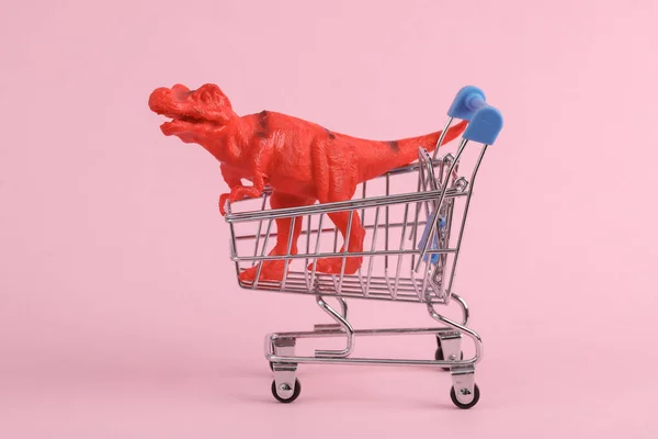Toy red dinosaur tyrannosaurus rex with supermarket trolley on pink background. Minimalism creative layout