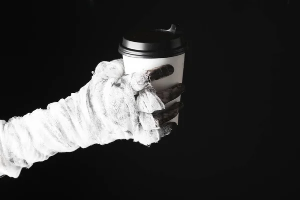 Mummy\'s hand wrapped in bandage holds coffee cup isolated on black background. Halloween concept