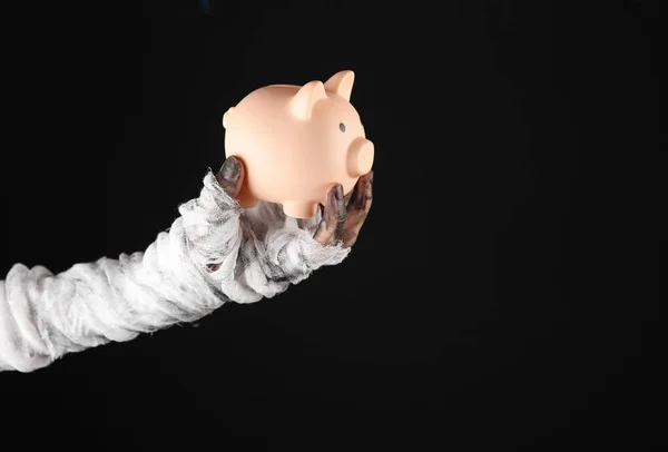 Mummy\'s hand wrapped in bandage holds piggy bank isolated on black background. Halloween concept