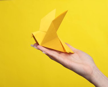 Sarı arka planda origami kağıt güvercini tutan kadın el.