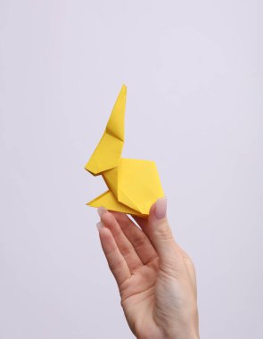 Kadın eli gri arka planda sarı origami tavşanı tutuyor. Hobi, yaratıcılık