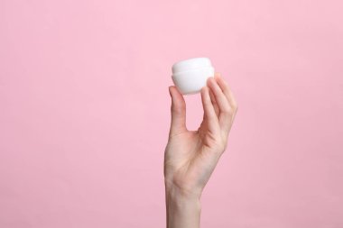 Kadının eli pembe arka planda beyaz krema kavanozunu tutuyor.