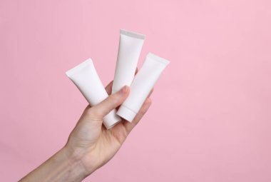 Kadının eli gri arka planda beyaz krem tüplerini tutuyor.