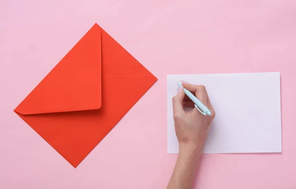 Mão Feminina Escreve Uma Carta Fundo Rosa Com Envelope Vermelho — Fotografia de Stock