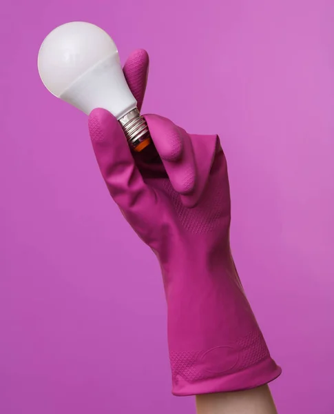 Hand in purple rubber cleaning glove holding led light bulb on a purple background. House cleaning and housekeeping concept