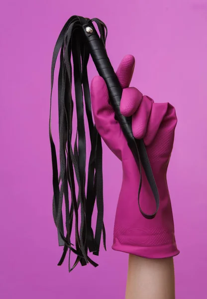 Hand in purple rubber cleaning glove holding sex shop leather whip on purple background.