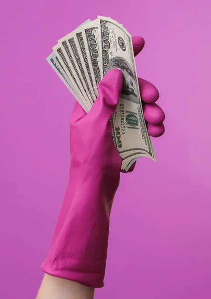 Hand in purple rubber cleaning glove holding dollar bills on purple background. House cleaning and housekeeping concept