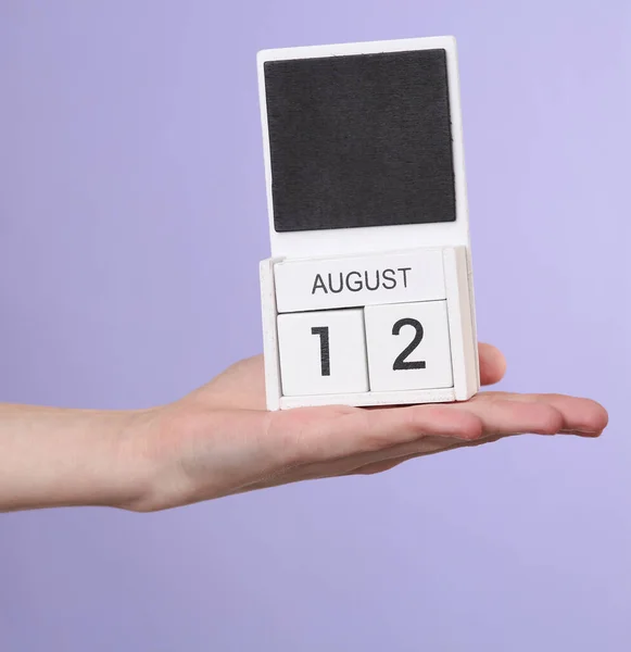 Woman\'s hand holds wooden calendar with date august 12 on pastel purple background