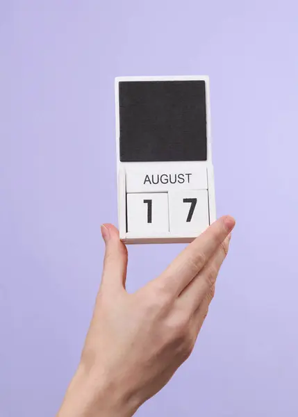 Woman\'s hand holds wooden calendar with date august 17 on pastel purple background