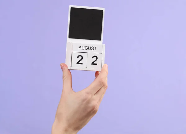 Woman\'s hand holds wooden calendar with date august 22 on pastel purple background
