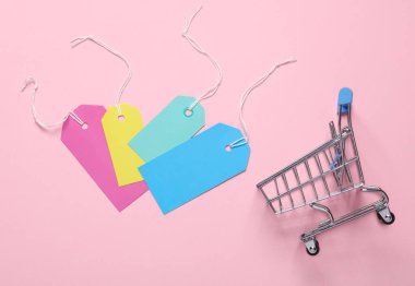 Pembe arka planda bir sürü boş fiyat etiketi olan mini alışveriş arabası. Satış, alışveriş konsepti. Üst görünüm