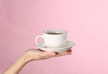 Kadın eli, pembe arka planda fincan tabağı olan beyaz bir seramik bardağı tutuyor.