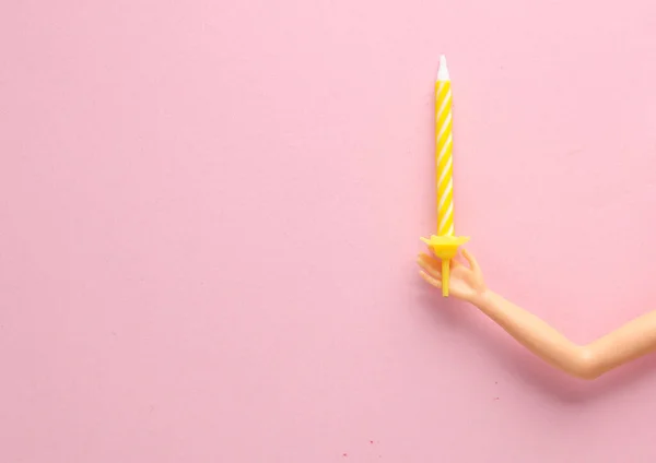 The doll\'s hand holds birthday candle on pink background. Minimal layout