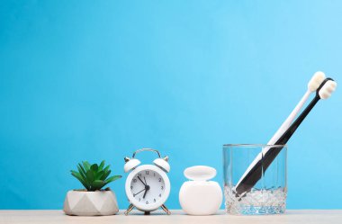 Diş bakımı. Bardakta iki diş fırçası, diş ipi, çalar saat ve masada bir bitki, mavi arka plan.