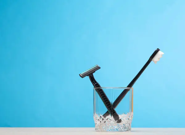 Disposable razor and toothbrush in a glass on the table, blue background. Daily routine, dental care and shaving