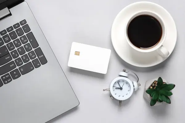 Laptop, White empty bank card with chip, cup of coffee, plant and an alarm clock on gray background. Flat lay business concept. Online shopping. Top view