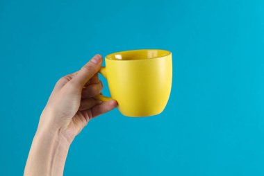 Kadının eli mavi arka planda sarı bir seramik bardak tutuyor.