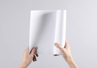 Kadın elleri, gri arka planda beyaz sayfaları olan boş bir dergi modeli tutuyor.