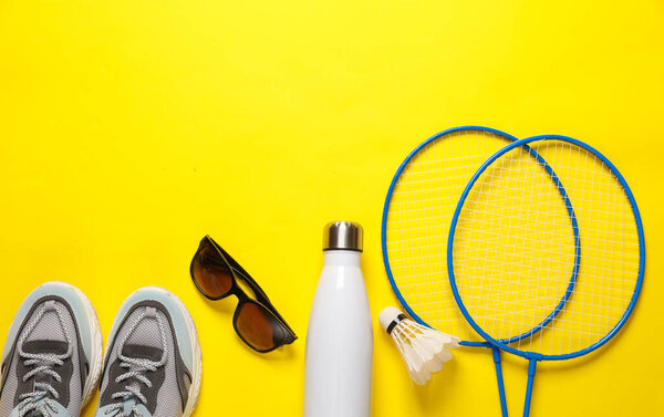 Badbintion racket with a shuttlecock and sneakers, sunglasses, thermos bottle on yellow background. Leisure. Top view