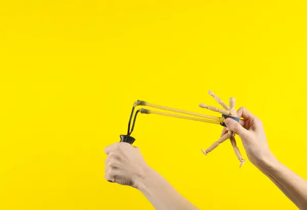 Hands holding slingshot with wooden puppet on a yellow background
