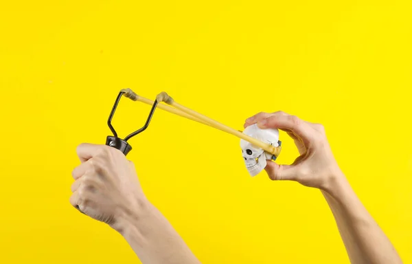 Hands holding slingshot with toy skull on a yellow background. Halloween theme, trick or treat