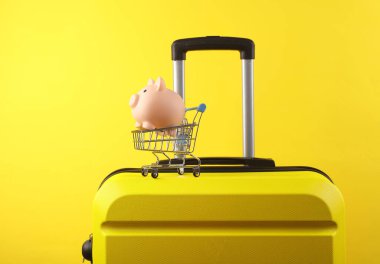 Sarı arka planda küçük bir süpermarket tramvayında domuz kumbarası olan sarı plastik bir valiz. Tatil alışverişi.
