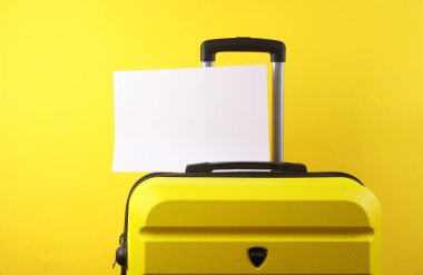 Sarı plastik bir valiz ve sarı arka planda boş bir kağıt. Turizm, tatil, taşınma konsepti. Boşluğu kopyala