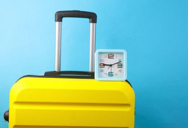 Sarı plastik valiz ve mavi arka plandaki saat. Turizm, tatil kavramı
