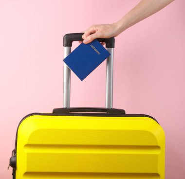 El ele tutuşan sarı plastik valiz sapı ve pembe arka planda pasaport. Turizm, tatil kavramı