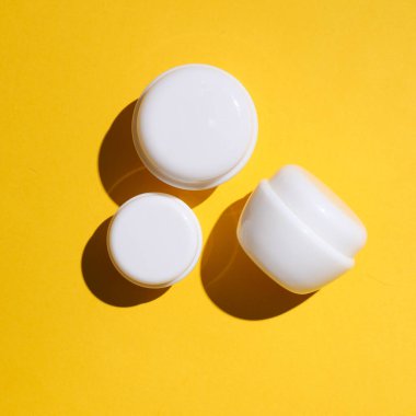 Sarı arka planda gölgeli üç beyaz krema kavanozu. Yaratıcı güzellik düzeni. Üst görünüm.